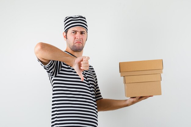 Young male holding cardboard boxes, showing thumb down in t-shirt, hat and looking discontent. front view.