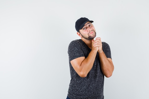 Young male holding clasped hands in praying gesture in t-shirt and cap front view.