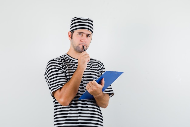 Giovane maschio che tiene appunti, penna in t-shirt, cappello e guardando pensieroso, vista frontale.