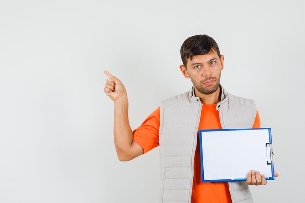 Young male holding clipboard, pencil, pointing left up in t-shirt, jacket front view.