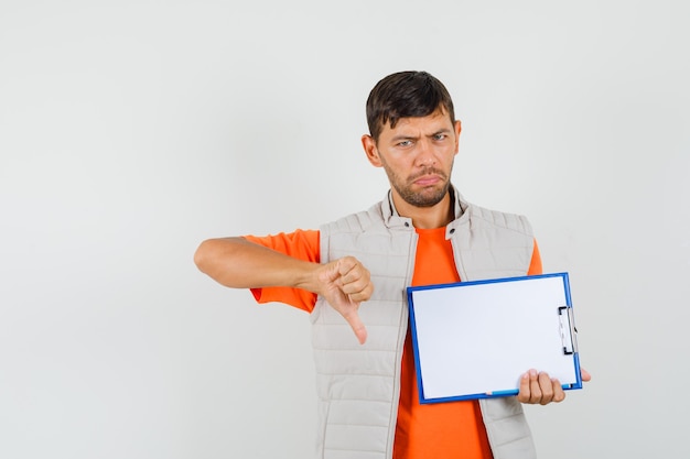 Young male holding clipboard, pencil, showing thumb down in t-shirt, jacket and looking upset , front view.