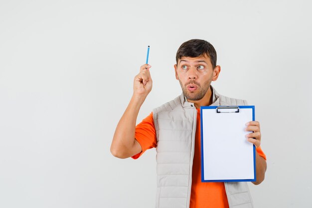 Молодой мужчина держит доску сзажимом для бумаги и карандаш в футболке, куртке и смотрит вдумчиво. передний план.