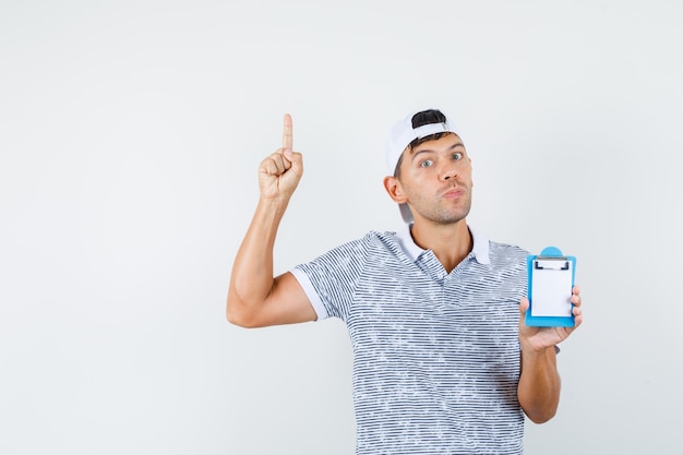 Young male holding clipboard, pointing up in t-shirt and cap and looking focused