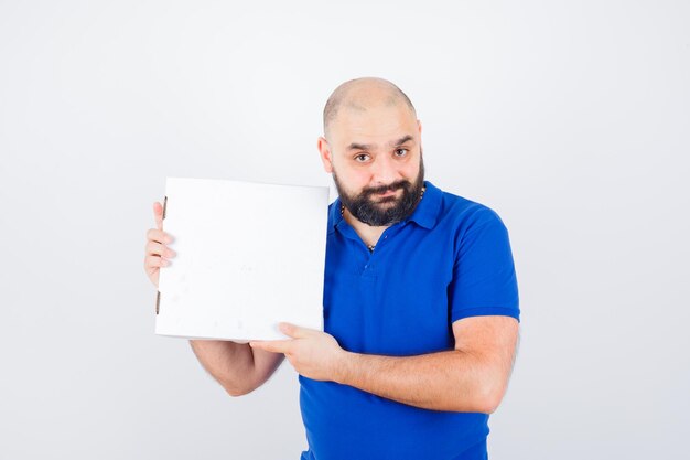 Young male holding closed pizza box in t-shirt and looking happy , front view.