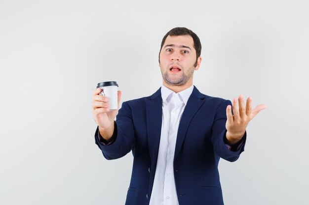 Young male holding cup of coffee in shirt, jacket and looking confused