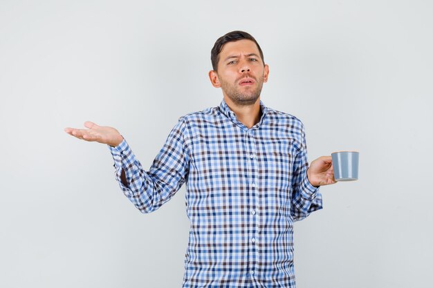 Молодой мужчина держит чашку напитка в озадаченном жесте в клетчатой рубашке