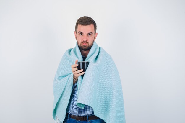 Young male holding cup while looking at camera in shirt, jeans, blanket and looking serious. front view.