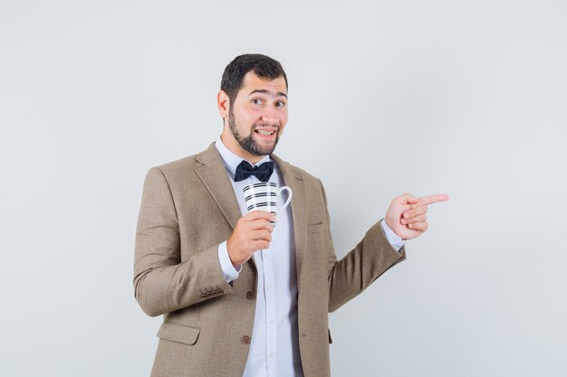 Young male holding cup while pointing to the side in suit and looking optimistic. front view.