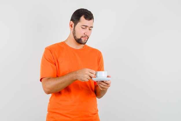 Young male holding cup with saucer in orange t-shirt and looking careful. front view.