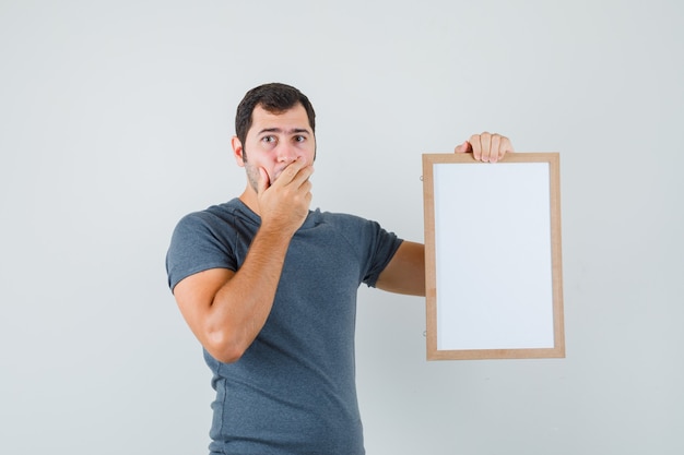 Free photo young male holding empty frame in grey t-shirt and looking anxious