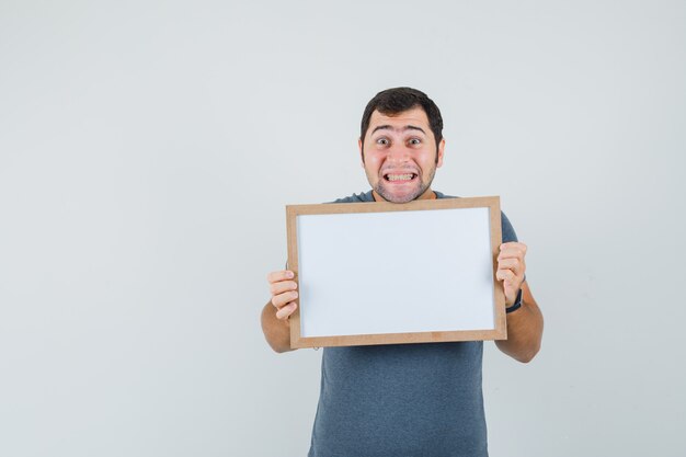 Young male holding empty frame in grey t-shirt and looking glad 