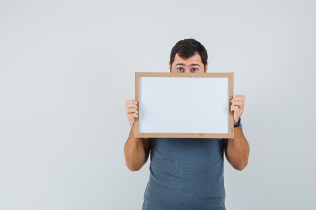 Young male holding empty frame in grey t-shirt and looking scared  