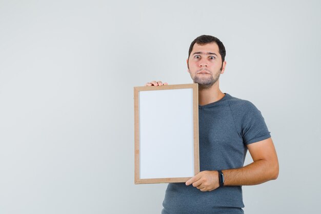 Young male holding empty frame in grey t-shirt and looking troubled  