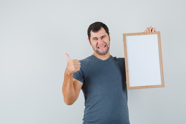 Young male holding empty frame showing thumb up in grey t-shirt and looking cheerful 