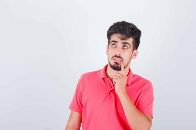 Young male holding finger on chin in pink t-shirt and looking pensive , front view.