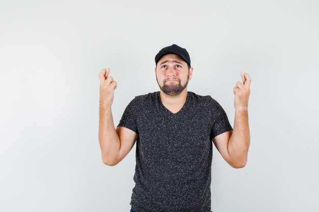 Young male holding fingers crossed in t-shirt and cap and looking hopeful