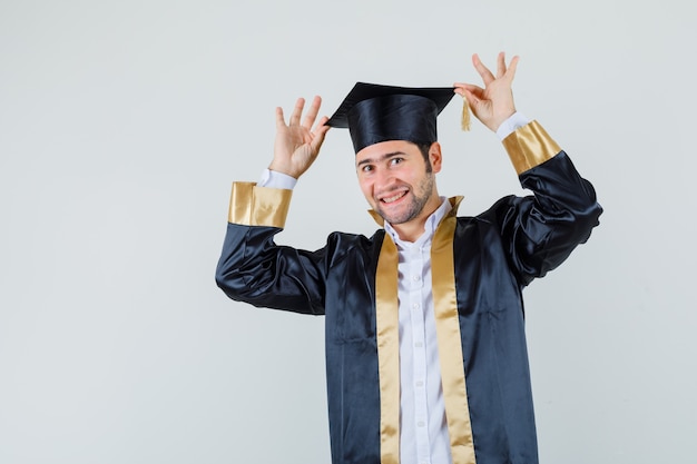 卒業式の制服を着た彼の帽子に指を持って、陽気に見える若い男性、正面図。