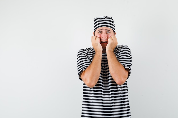 Free photo young male holding fists on cheeks in t-shirt, hat and looking anxious , front view.