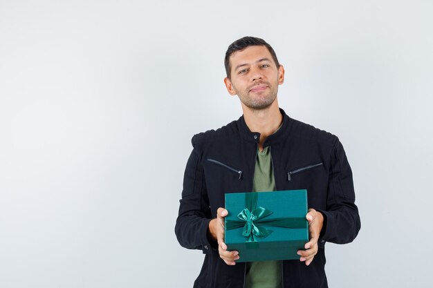 Young male holding gift box in t-shirt, jacket and looking glad , front view.