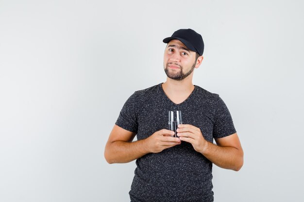 Young male holding glass of water in t-shirt and cap and looking pleased 