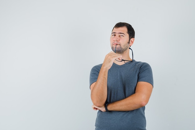 Young male holding glasses in grey t-shirt and looking thoughtful 