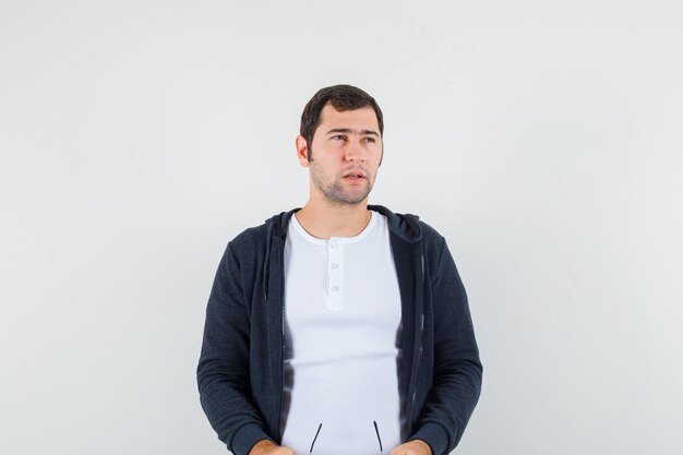 Young male holding glasses in t-shirt, jacket and looking pensive , front view.