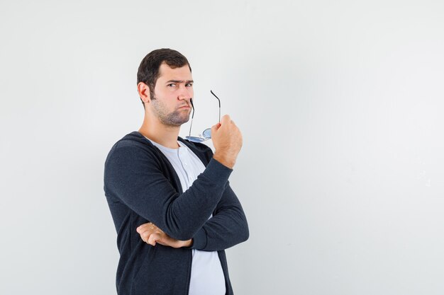 Young male holding glasses in t-shirt, jacket and looking thoughtful. front view.