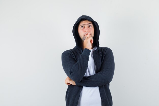 Young male holding hand on chin in jacket, t-shirt and looking pensive. front view.