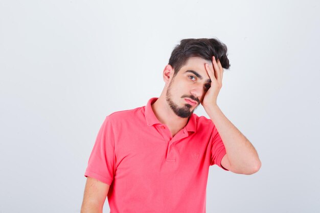 Young male holding hand on face in pink t-shirt and looking pensive. front view.