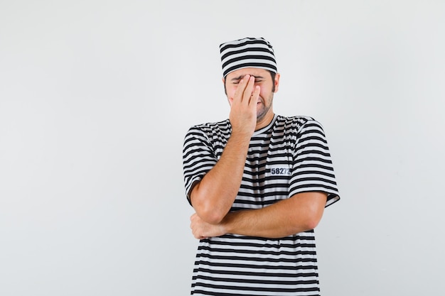 Young male holding hand on face while crying in t-shirt, hat and looking depressed. front view.