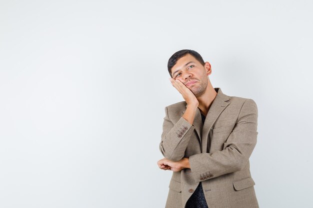 Young male holding hand on his face in grayish brown jacket and looking dreamy , front view.