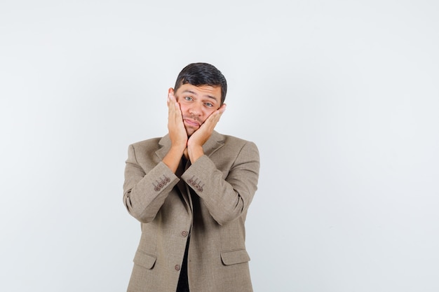 Young male holding hand on his face in grayish brown jacket and looking mournful , front view.