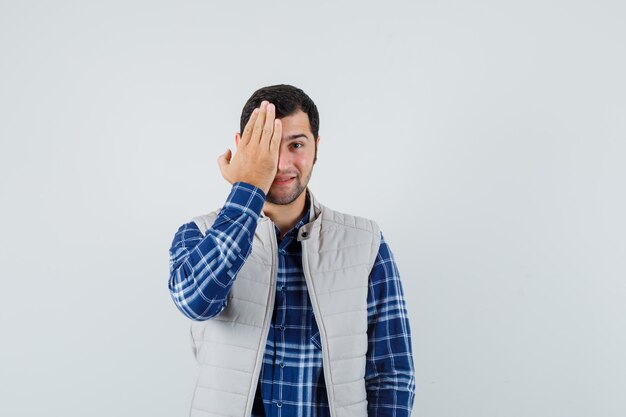 Young male holding hand on his face in shirt,sleeveless jacket and looking joyful. front view.