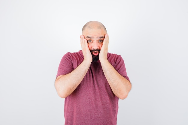 Young male holding hands on cheeks in pink t-shirt and looking scared , front view.