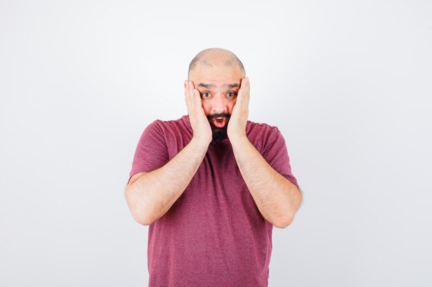 Young male holding hands on cheeks in pink t-shirt and looking scared , front view.