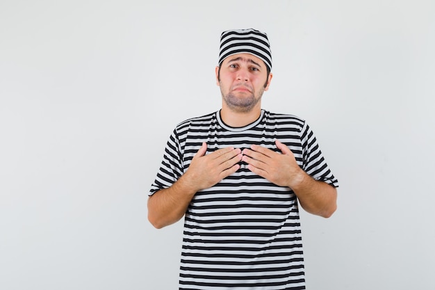 Young male holding hands on chest in t-shirt, hat and looking mournful , front view.