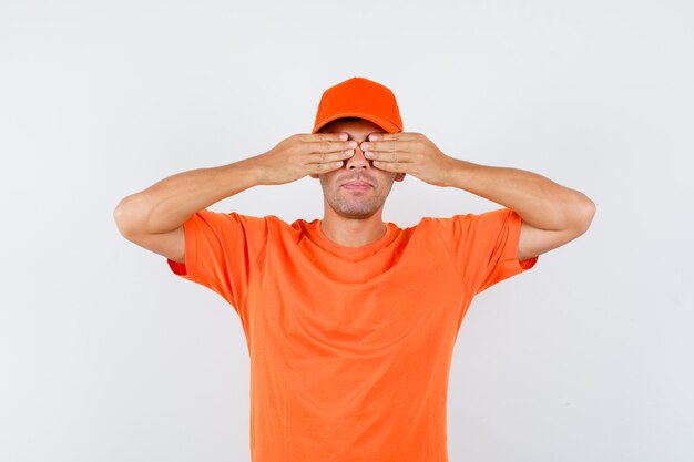 Young male holding hands on eyes in orange t-shirt and cap front view.