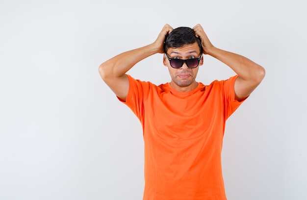 Young male holding hands on head in orange t-shirt and looking puzzled