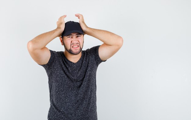 Young male holding hands on head in t-shirt and cap and looking regretful