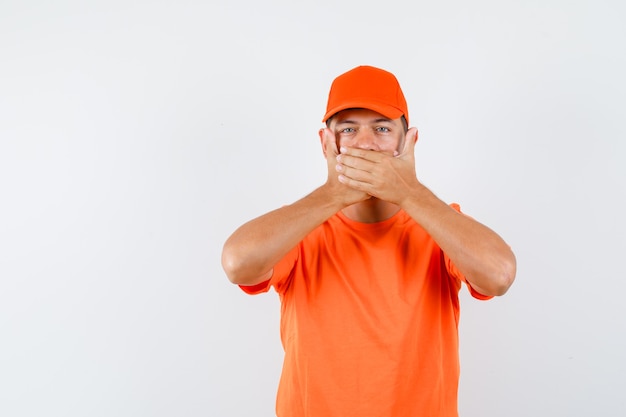 Young male holding hands on mouth in orange t-shirt and cap and looking scared