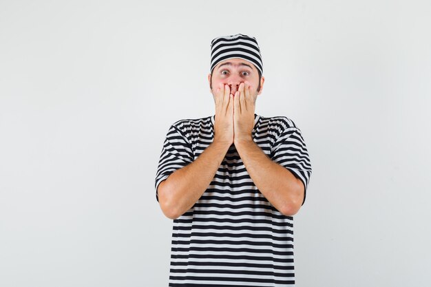 Young male holding hands on mouth in t-shirt, hat and looking scared. front view.