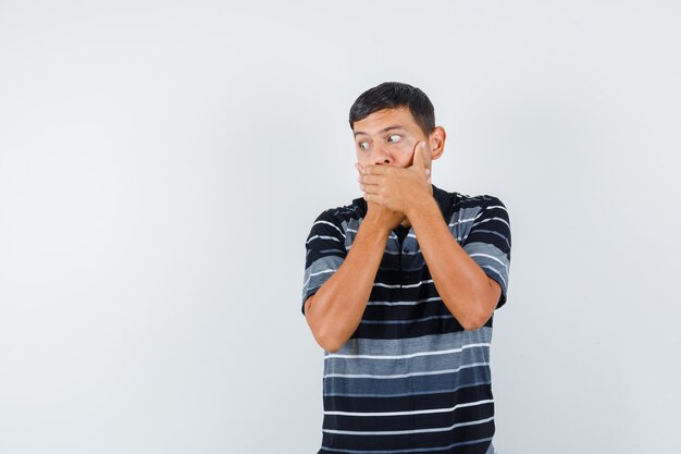 Young male holding hands on mouth in t-shirt and looking scared , front view.