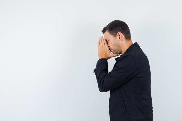 Young male holding hands in praying gesture in black jacket and looking hopeful. .