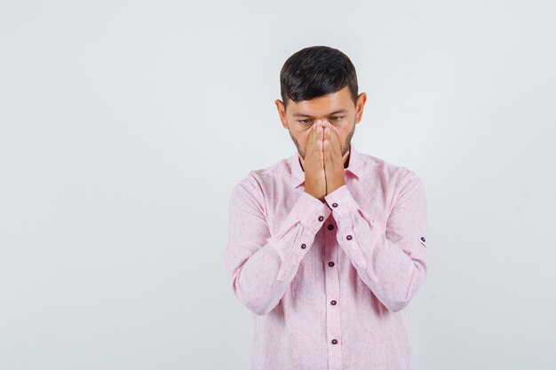 Young male holding hands in praying gesture in pink shirt and looking hopeful , front view.