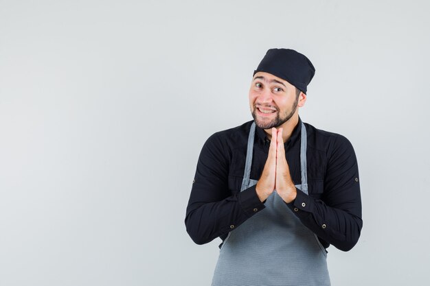 Young male holding hands in praying gesture in shirt, apron and looking joyful , front view.