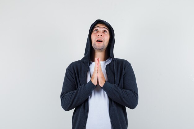 Young male holding hands in praying gesture in t-shirt, jacket and looking hopeful. front view.