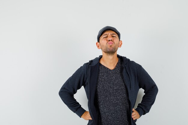 Young male holding hands on waist, pouting lips in t-shirt, jacket, cap , front view.