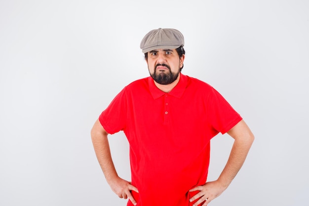 Young male holding hands on waist in red t-shirt,cap and looking displeased , front view.