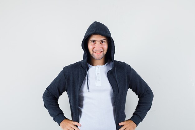 Young male holding hands on waist in t-shirt, jacket and looking confident. front view.