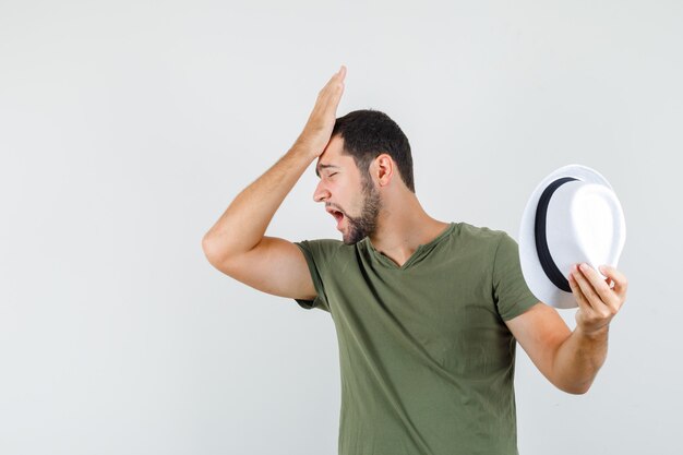 Young male holding hat with hand on forehead in green t-shirt and looking forgetful